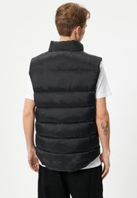 Schwarze gepolsterte Weste mit hohem Kragen, ausgestattet mit horizontaler Steppung und glatter Textur. Über einem weißen Hemd getragen, von hinten dargestellt.