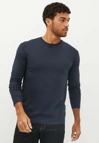 Hosszú ujjú sötétkék póló sima anyagból, kerek nyakkivágással és fitted szabással. Sötét farmernadrággal párosítva.