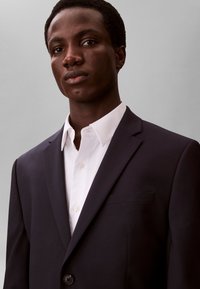 Ung mand med kort hår iført en mørk jakkesætjakke over en hvid skjorte med knapper, vendt fremad mod en almindelig grå baggrund.