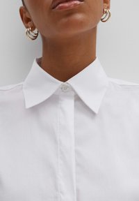Camicia bianca con colletto puntuto, texture liscia e chiusura con un bottone al collo. Orecchini a cerchio dorati visibili.