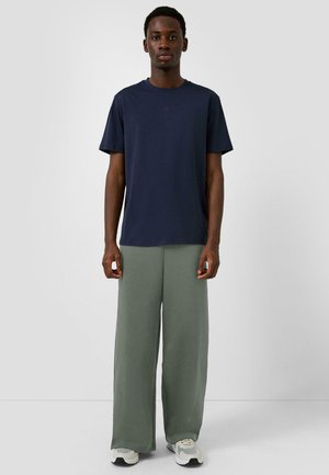 Homme debout portant un t-shirt bleu marine à manches courtes, un pantalon ample vert olive et des baskets blanches, sur un fond uni.