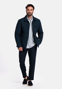 Gequiltede marineblauwe jas met een kraag, knopen aan de voorkant, zijzakken, gedragen over een gestreept shirt en donkere broek, aangevuld met zwarte schoenen.