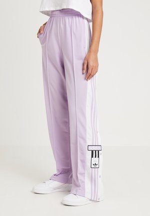 Personne portant un pantalon de jogging Adidas lavande avec des bandes blanches sur les côtés et des écussons, assorti à des baskets blanches et un top court blanc.