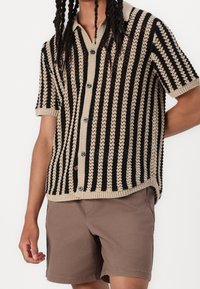 Korte mouw gebreide cardigan met zwarte en beige verticale strepen, knopen aan de voorkant en textuurpatronen. Gecombineerd met een paar bruine korte broeken.