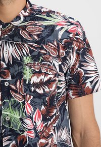 Košile s krátkým rukávem na knoflíky s tmavě modrým pozadím a živými tropickými vzory listů v hnědé, zelené, bílé a červené barvě.