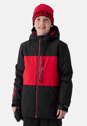 Garçon portant une veste d'hiver noire et rouge avec le logo "Spyder" et un bonnet rouge assorti, souriant sur un fond uni.