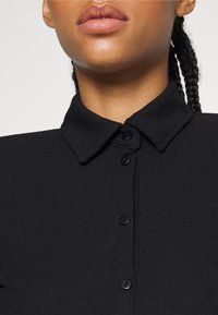 Lähikuva henkilöstä, joka on pukeutunut mustaan ribbipaitaan, jossa on kaulus, ja näkyvissä on kolme pitkää letkua olkapään takana.