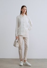 Λευκό πουκάμισο με κουμπιά και απαλό ύφασμα, συνδυασμένο με ανοιχτό μπεζ στενά παντελόνια και λευκά αθλητικά παπούτσια· κρατώντας μια μικρή μπεζ τσάντα χειρός.