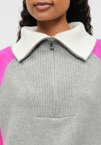 Sweater en tricot gris avec un col blanc côtelé, doté d'une fermeture éclair quart et de manches raglan roses. Texture simple avec un design épuré.