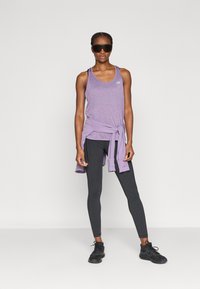 Μωβ αθλητικό τοπ με δέσιμο, συνδυασμένο με μαύρο κολάν και μαύρα αθλητικά παπούτσια. Λείο ύφασμα, με λεπτομέρεια μικρού λογότυπου στο τοπ.