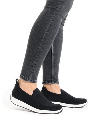 Beneden van een persoon met strakzittende donkergrijze spijkerbroek en zwarte athletykschoenen zonder veters met witte zolen, op een witte achtergrond.