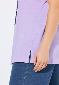 Camisa de lavanda con aberturas laterales, de tejido ligero, llevada sobre denim oscuro. Primer plano de una mano descansando en el dobladillo de la camisa.