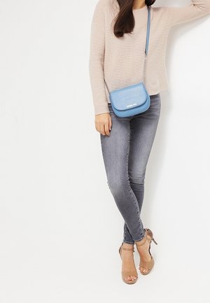 Donna con maglione beige e jeans skinny grigi, porta una piccola borsa a tracolla blu, in piedi con le gambe incrociate e tacchi beige.