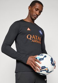 Μαύρο αθλητικό t-shirt με μακριά μανίκια και πορτοκαλί κείμενο "QATAR AIRWAYS" και λογότυπο; το μοντέλο κρατάει μια πολύχρωμη μπάλα ποδοσφαίρου.