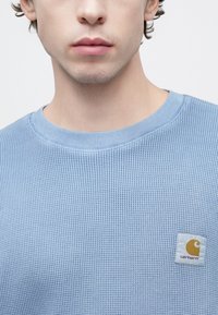 Ανοιχτό μπλε πουλόβερ πλεκτό σε ύφανση βαφλοειδούς μοτίβου με στρογγυλή λαιμόκοψη και τετράγωνο πλαϊνό σχέδιο με το λογότυπο Carhartt σε κίτρινο και πορτοκαλί στο αριστερό στήθος.