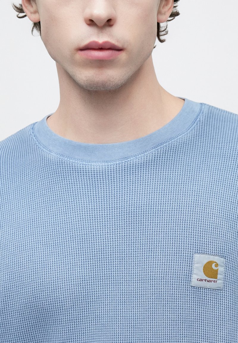 Light blue waffle-knit sweater featuring a crew neck and a square patch with the Carhartt logo in yellow and orange on the left chest.