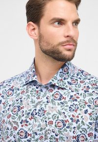 Camicia con bottoni caratterizzata da un tessuto azzurro chiaro con un motivo floreale in rosso, verde e blu navy. Colletto standard e dettagli dei bottoni ben visibili.