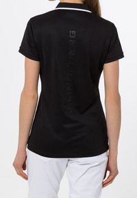 Černé polo tričko s jemným leskem, krátkými rukávy a kontrastním bílým límečkem. Na zádech je vyražené logo značky.