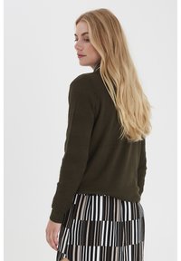 Olivgrüner gestrickter Pullover mit geripptem Muster an den Ärmeln, kombiniert mit einem schwarz-weißen gestreiften Rock mit unterschiedlichen Breiten.