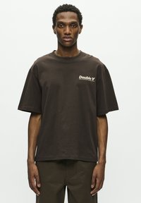 Hombre con cabello rizado corto, vistiendo una camiseta suelta de color marrón oscuro con el texto "Double A" en el pecho, de pie contra un fondo blanco liso.