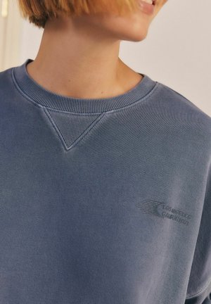 Niebieska bluza z ribbowym okrągłym dekoltem, z charakterystycznym haftem w kształcie trójkąta oraz małym nadrukowanym logo na klatce piersiowej w kontrastującym kolorze.