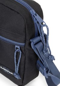 Černá látková crossbody taška s modrými zipy a popruhy, nastavitelným ramenním popruhem a logem značky na přední straně.