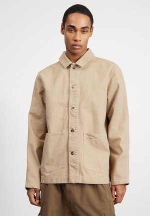Μπεζ καμβάς εργασίας μπουφάν με γιακά, μπροστινά κουμπιά και δύο τσέπες επένδυσης. Διαθέτει διακριτική ραφή και ελαφριά υφή.