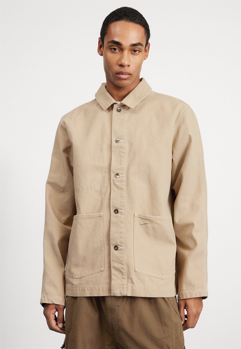 Chaqueta de trabajo de lienzo beige con cuello, botones frontales y dos bolsillos de parche. Presenta costuras sutiles y una leve textura.