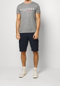 Man i en grå Hilfiger-t-shirt, marinblå cargoshorts, vita strumpor och vita hög-top sneakers, som står mot en enkel bakgrund.
