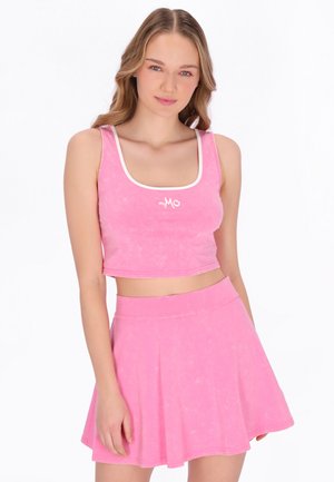 Fiatal nő rózsaszín crop topot visel fehér szegéllyel és hozzá illő, bővülő szoknyát, fehér háttér előtt pózolva.