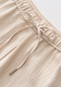 Tessuto beige chiaro con un motivo a righe texturizzato. Presenta una vita elastica con cordoncino legato e un dettaglio in metallo argentato alla fine del cordoncino.