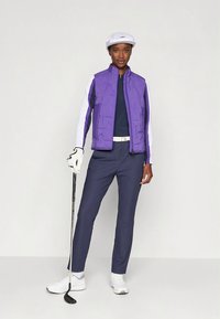 Fioletowa pikowana kamizelka na granatowej bluzce, do tego granatowe spodnie. Biała czapka i rękawiczki. W ręku kij do golfa. Całość dopełniają białe tenisówki.