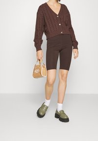 Mujer con un cárdigan marrón de punto de cable y pantalones cortos de ciclista a juego, sosteniendo un bolso color beige, con calcetas blancas y mocasines gruesos color verde oliva.