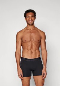 Boxer shorts pretos com uma cintura suave, apresentando o logótipo "Levis" em destaque, usados por um modelo masculino sem camisete.