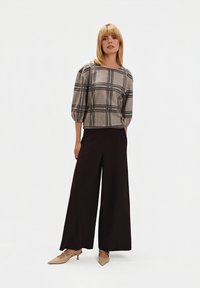 Szary i czarny top w kratę z rękawami do trzech czwarteks, w połączeniu z szerokimi czarnymi spodniami i beżowymi szpilkami na odkrytej pięcie. Gładkie białe tło.