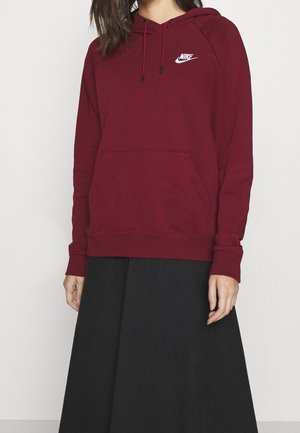 Frau trägt einen bordeauxroten Nike-Hoodie mit Fronttasche und Kordelzügen, kombiniert mit einem schwarzen Rock, vor einem einfarbigen Hintergrund.