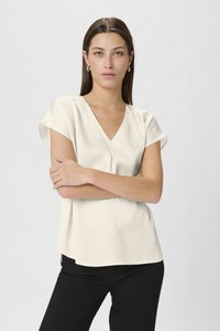 V-neck, short-sleeve blouse in light cream, made of textured fabric; paired with black trousers, model crossing arms against a neutral background.