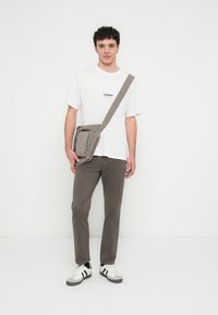 Pilka per petį dėvima rankinė su priekinė kišenė su užtrauktuku, dėvima ant balto marškinėlių, derinama su rudos spalvos kelnėmis ir baltais sportbačiais su juodais akcentais.