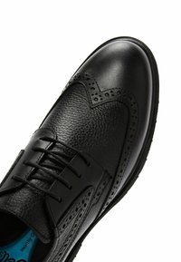 Chaussure habillée en cuir noir avec surface texturée, design à bouts ailés, détails brogues le long du bord, et lacets. La semelle est plate et robuste.