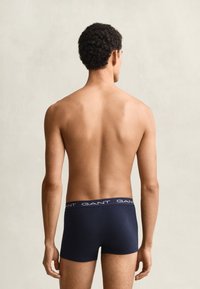 Mann mit kurzen lockigen Haaren, von hinten gesehen, trägt marineblaue GANT Boxershorts vor schlichtem hellem Hintergrund.