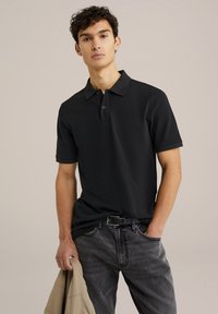 Mustat polo paidat, joissa on klassinen kaulus, lyhyet hihat ja kaksipainike malli, yhdistettyinä harmaisiin farkkuihin ja vaaleanbeigeen takkiin.