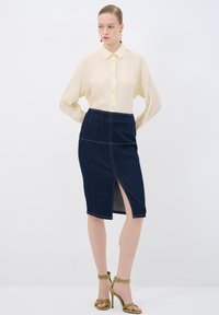 Camisa amarilla claro de botones y mangas largas combinada con una falda lápiz de mezclilla oscura con una abertura delantera, complementada con tacones verde olivo.
