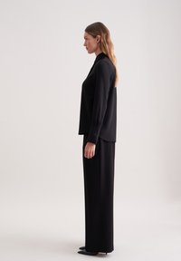 Blusa nera a maniche lunghe con vestibilità ampia e polsini abbottonati, abbinata a pantaloni neri a gamba larga. Tessuto morbido e design sartoriale.