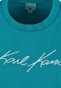 Turkoza krāsa kokvilnas t-krekls ar noapaļotu kaklu un izšūtu balto tekstu "Karl" rūtiņā uz priekšpuses. Mīksts audums, ikdienišķa forma.