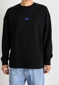 Svart sweatshirt tillverkad av mjukt tyg, med en blå "HUGO" logotyp på framsidan, med långa ärmar och avslappnad passform.