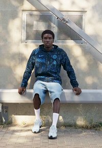 Nike hættejakke i mørkeblå med abstrakt mønster, parret med lyse denimshorts og hvide sokker, siddende på en bænk ude.