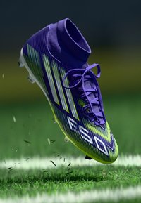 Ghete de fotbal violet și verde, cu un guler de tip sosetă, suprafață texturată, trei dungi albe și un logo F50, suspendate deasupra ierbii.