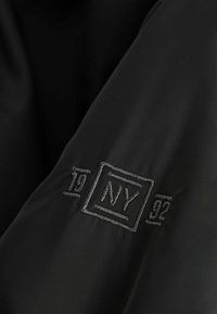 Czarny materiał z wyraźnym haftowanym logo z napisem "NY" i rokiem "92" w prostokątnym obramowaniu. Gładka tekstura, minimalistyczny design.