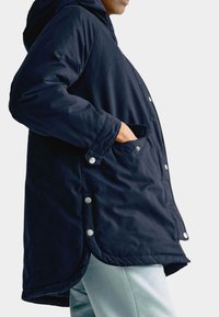 Námorně modrá parka s kapucí, s patentkami po stranách, bočními kapsami a mírně zakřiveným spodním lemem. Materiál se jeví jako hladký a strukturovaný.