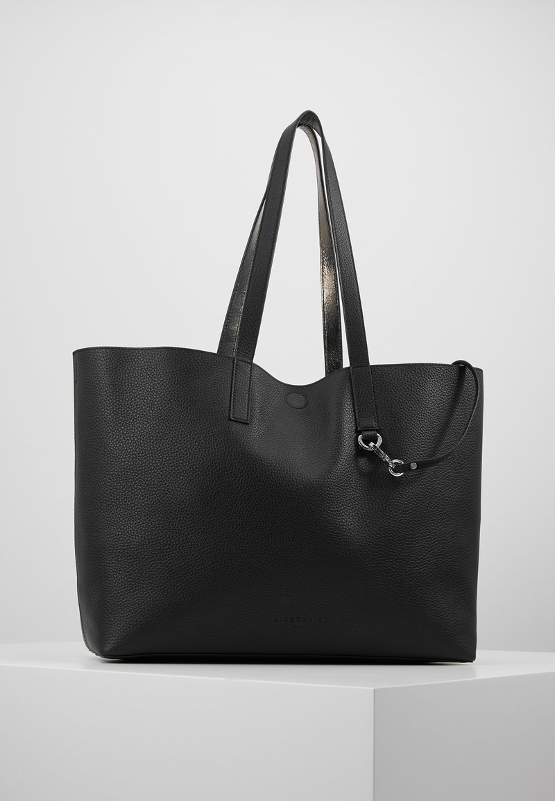 Sac cabas en cuir noir texturé avec deux longues anses et une petite sangle attachée avec une boucle métallique, posé sur un piédestal blanc.